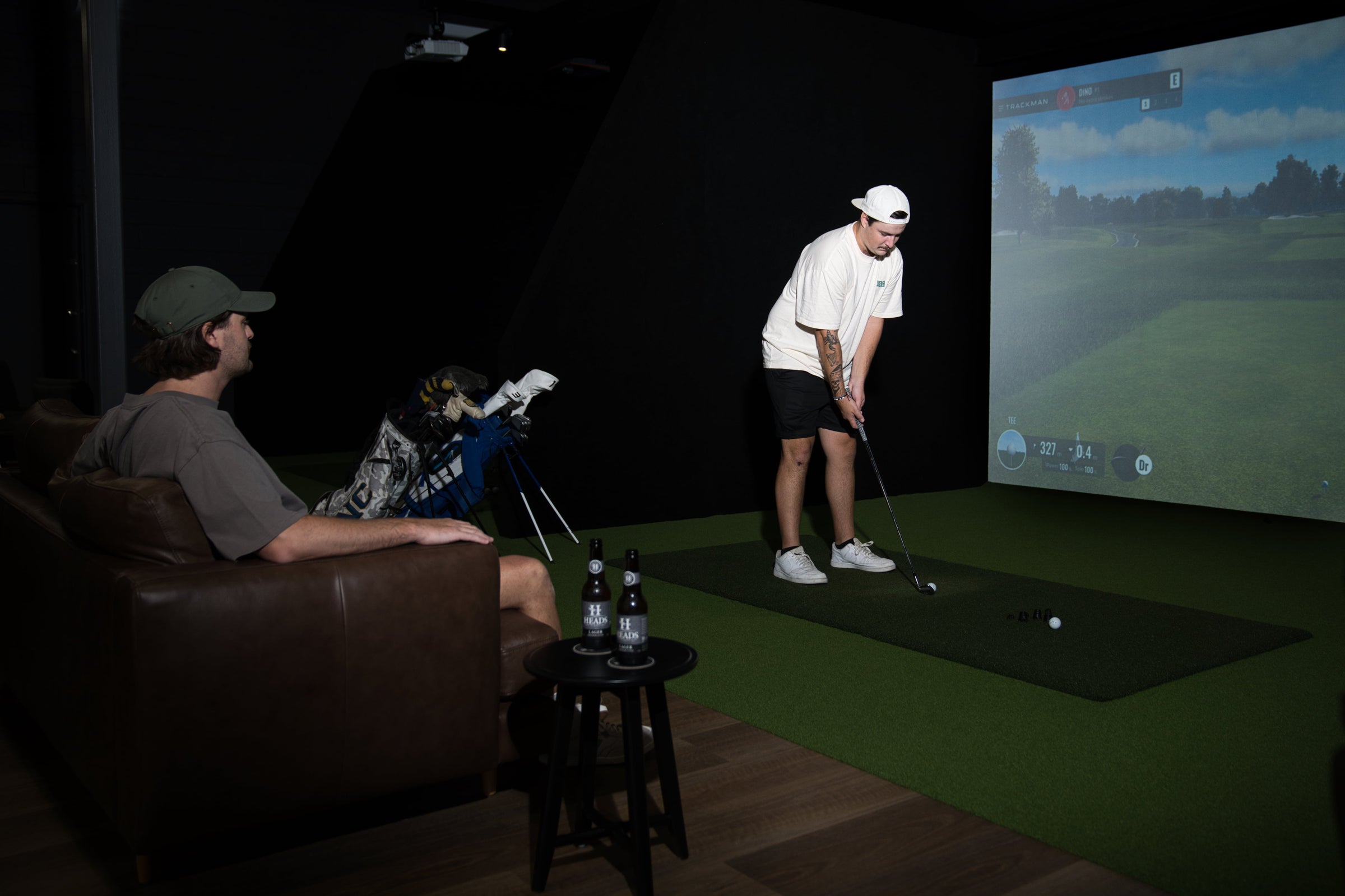 Golfers using a TrackMan indoor golf simulator at Noosa Golf Co. on the Sunshine Coast. One player tees off while another relaxes nearby with golf gear and local drinks in view. A projected course is displayed in the background.